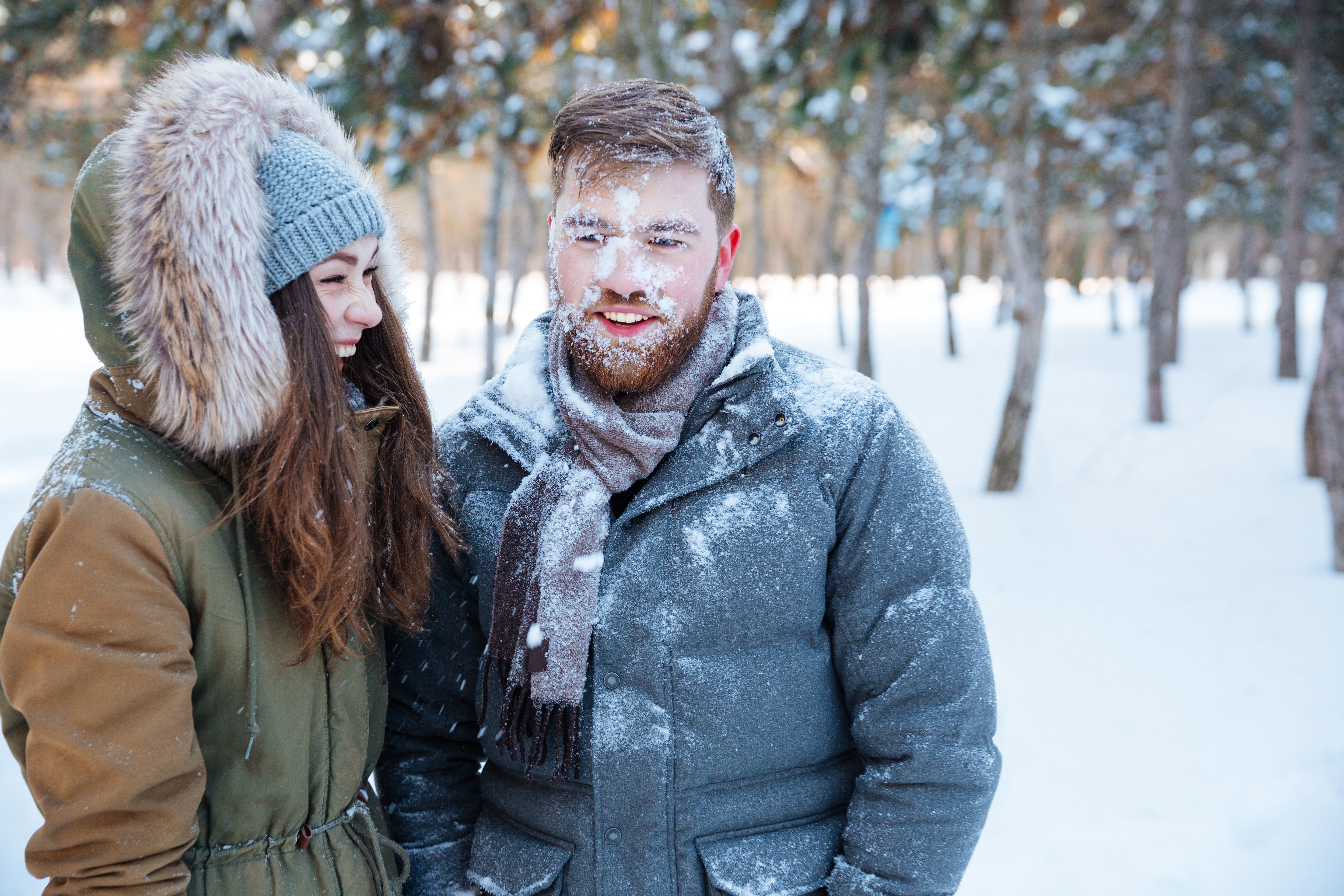 Couple standing in a snowy forest with snow on their faces and coats, illustrating the effects of winter weather on skin.